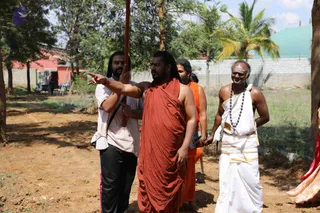 A photograph of an outdoor construction site among trees and dirt ground. A man in a long rust-red wrap and rudraksha-style beads, with long dark hair, extends his arm and points toward the left while a man in a striped pink-and-white shirt and black trousers holds a tall red pole upright beside him. Behind them a man in an orange garment stands near a hand-held device, and on the right a bald, smiling man in a white dhoti with gold borders and a long beaded necklace stands with his hands clasped. A low cinderblock wall, palm trees and a green-roofed structure appear in the background, with a red building to the left.