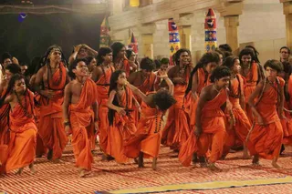 A group of young dancers in saffron-orange dhoti-style garments and rudraksha malas performs energetically on a striped red and cream mat in a pillared hall. Several dancers have long braided hair with beaded strands, and some wear beaded necklaces and bangles. Behind them stand carved stone pillars topped with tall decorative poles painted with blue, yellow and red geometric motifs, and a few spectators watch from the edges. The dancers are mid-movement, arms and legs flung out, with expressive faces.