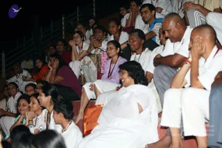 Photo of a large seated audience at an evening satsang, packed onto tiered concrete steps in a dark venue. Most attendees wear white clothing with red lanyard-style cords or beaded necklaces. In the foreground a woman in a white sari sits beside an orange-and-gold cloth bag. Behind her a woman in a lilac top and white pants looks ahead, and a man in a grey shirt and dark trousers sits at right. Several people have hands raised to their faces or clasped in attention. No one at the front of the stage is visible, and no a person candidate appears in this frame.