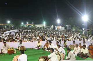 Night-time outdoor gathering under bright floodlights, with a very large crowd seated on green mats across an open ground. In the foreground, several men in white dhotis and shirts sit playing drums, including a large barrel drum with a red and yellow body, and a long wind instrument. Behind them rows of attendees in white and cream clothing fill the space, with some saffron-robed figures at the right edge. Strings of lights and a tented stage area are visible in the background.