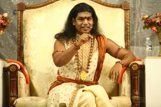 A seated man on an ornate cream and gold throne-style chair speaks while gesturing with his right hand. He has long dark hair, a red bindi on his forehead, and a small headset microphone. He wears a cream and gold patterned robe with an orange shawl draped over one shoulder and tied at the waist, multiple rudraksha malas and bracelets, and a large gold pendant. Flowers in a basket and a water bottle sit at the right edge against a glittering grey backdrop.