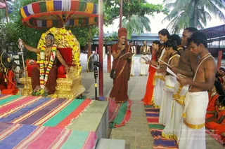 Outdoor ceremony on a paved courtyard under a palm tree and a multicolored canopy. A man seated on a gilded throne with red cushions, wearing a crown, a red-brown garment and garlands of yellow and white flowers, raises a silver ceremonial staff. In front of him a woman in a rust-red saree holds a microphone and reads from papers, while a row of men and women in white dhotis and orange saris, some bare-chested, stand holding printed pages. Striped rugs cover the ground, and a standing fan and a microphone stand are at the side. a person cues are limited here, so the seated figure is identified only as the seated presiding figure on the throne.