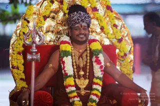 A photograph of a smiling man seated on a red-cushioned golden throne with a tall ornate golden backrest, wearing a leopard-print headwrap and a long-sleeved maroon-orange robe. He wears a heavy gold necklace with a large pendant, a rudraksha-style bead mala, and gold armbands, and a sweeping garland of yellow marigolds, white flowers, red roses and green leaves drapes over his shoulders and arms. A silver trident stands at his left side. A blurred attendant in white stands at the right edge.