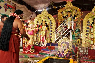 A long-haired man in a rust-red dhoti and red shawl with a gold armlet stands at the left foreground, facing a line of decorated deity murtis under a tent canopy. He is offering flowers and petals near a pink Ganesha murti in gold dress. Behind them, a golden-framed Devi Durga murti with multiple arms holds a trident and weapons, with a green lion-like vahana below. Garlands of marigolds hang on the murtis, and brass oil lamps and petal-strewn cloth surround a yellow-edged pit with a floral garland. A white-clad attendant is partially visible at the far left edge. A world map hangs on the wall behind.