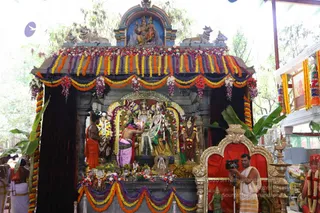 A photograph of a decorated shrine with a deity murti of Shiva and Parvati, adorned with marigold and multicoloured flower garlands in orange, yellow, purple and pink. A priest in a pink dhoti bends to make offerings at the central image, while another attendant in an orange cloth stands beside him. A cameraman in a white dhoti films the ritual from the right, beside a gilded throne draped in red. Above the shrine, a painted panel shows Shiva and Parvati seated with nandi bulls. Oil lamps and flower offerings sit on the platform.
