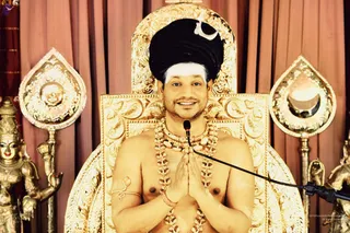 A bare-chested man with a tall black turban and white urdhva tilak sits on an ornate gold throne with carved scrollwork and a crescent motif, pressing his palms together in anjali. He wears layered gold and bead necklaces and a gold armlet, with a microphone on a stand beside him. Gold deity statues flank the throne against draped rust-coloured curtains. Whether he is the person named on the page cannot be confirmed from appearance alone, so the identity is marked unsure.