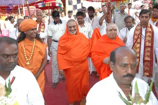 A procession on a red carpet under decorated canopies with colourful tassels. At the centre, a smiling bearded man in a saffron-orange robe, drawn over his head, walks forward. Beside him, a bald man with white tripundra marks on his forehead wears an orange shawl. On the left, a person with long dark hair, an orange head wrap and an orange robe wears a gold necklace with a pendant. Behind them, men in white shirts and dhotis wear red-and-gold bordered shawls, some carrying long poles. In the foreground, a man in white with a red sash and a bald man in white are seen partially, with white flowers and green leaves at the lower left. Printed text at the lower right reads Sri Nithyananda Dhyanapeetam.