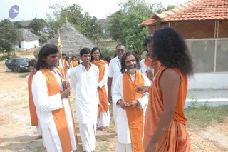 Outdoor photo on a dusty path beside a thatched hut, a tiled-roof building and a parked dark car. A young man in white kurta and dhoti with an orange shawl holds a tall gold-tipped staff, and the group of men in white and orange shawls walks and talks in a loose cluster. In the centre, a bearded man with long dark hair wears a white kurta and orange shawl and smiles with hands clasped. On the right, a long-haired man in an orange dhoti and orange shawl with a rudraksha mala faces him, gesturing with one hand. Several other men in white and orange stand behind, with trees and a village backdrop.