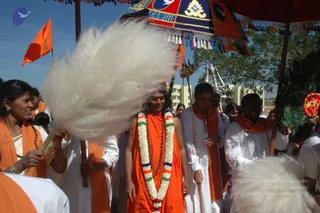 A festival procession on a sunny day under a red and blue canopy embroidered with a gold emblem and the word Peetam. In the centre, a young man in an orange saffron robe with a multicolour-and-white flower garland and rudraksha beads walks smiling, with a large white feathery fan-like whisk behind him. To the right, a man in white with an orange shawl and glasses walks beside him, and another man in white holds a peacock-feather fan. On the left, a woman in an orange shawl holds a white chamara-style whisk, and orange flags and a trident-topped staff appear in the background. Several other devotees in white and orange stand nearby under trees and a yellow building.
