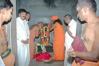 A temple sanctum with a dark stone Shiva lingam decorated with red and white flower garlands, a yellow marigold garland and a pink cloth offering at its base. A bare-chested elder in orange cloth bends forward in worship. Beside him, a person in orange robes with a saffron head wrap and long dark hair bows with hands raised to the head, and a man in an orange upper cloth stands at the right holding a maroon bag, wearing a rudraksha mala. Two men in white dhoti and shirt stand at the left and centre-right with hands folded in reverence. Grey stone walls surround the scene, with a flower-decorated shrine and devotional offerings.