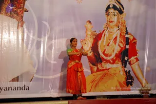 A female classical dancer in a red and orange silk sari with gold border and a green blouse stands on a stage in a striking pose, hands raised in a mudra near her chest. Behind her is a large printed banner of a goddess figure in gold and red garments, adorned with a crown, long braid, pearl garlands and gold jewellery, holding a lotus-like object. The banner edge shows partial lettering. A single dancer is the only physical person visible.
