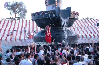 Outdoor photograph of a large black stone Shiva lingam fountain-basin with water pouring down its sides from the column above. A figure in a red garment with a white flower garland stands on the basin's platform amid scattered petals, arms spread, while water cascades around them. Below, a dense crowd of white-clad devotees, many with raised hands, stands in shallow water in front of a red-and-white striped stepped structure. A trident-like ritual object is visible at lower left. Trees and a small structure stand in the background under a pale sky.