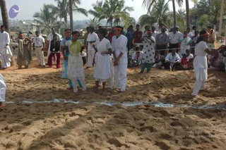 Outdoor photo of a traditional Indian game on a sandy field, with young women in white and orange headbands stepping across a marked line in the sand. Several women in white salwar-style outfits and a green-and-white sari stand together in the centre, one in a teal top. Behind a rope line, a crowd of spectators in white shirts, dark trousers and light dresses watches, with palm trees and buildings in the background. No identifiable a person figure is visible.