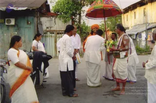 A photograph of a street scene with a group of people gathered on a paved road beside corrugated metal walls and a blue-painted building. A man in the centre wears an orange-saffron head wrap and orange-saffron garment over white dhoti, standing near a bearer holding a red, green and yellow decorated umbrella overhead. Another man on the right wears a white dhoti and shawl with a red-bordered cloth, bare-chested with white painted markings on his torso, holding a bouquet of yellow and pink flowers. Several men in white kurtas and white dhotis stand around, and a woman in a white sari with an orange border stands at the left edge beside a parked motorbike. Laundry hangs on a line at right. Visible printed copyright text appears at the lower right.