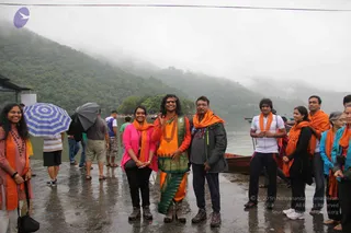 A group of people stands on a wet stone lakeside landing beside a misty, forested hillside and a moored wooden boat. In the centre, a man with long dark hair and a headband wears an orange top, a green-and-gold patterned wrap, a green shawl and a large beaded necklace, holding up two fingers in a peace sign. Around him, several people wear orange shawls; a woman in a pink cardigan and dark trousers stands at left, and a man in a dark jacket stands beside the central figure. On the left, a blue-and-white umbrella and a dark umbrella are held up in the drizzle. The people are visibly arranged for a group photo.