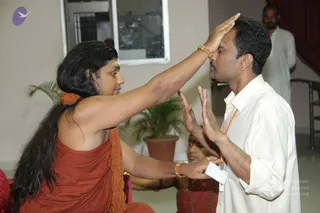A photograph of a seated man in a rust-orange robe with long black hair, a long orange-beaded mala around his neck and gold bracelets, pressing his right palm to the forehead of a man in a white shirt with an orange lanyard and an ID badge. The recipient stands with eyes closed and hands raised, while another orange-robed person's hands are visible at lower left. A potted plant, a window and a white wall form the background, with a standing man in white at the upper right. a person is the orange-robed figure seated with long hair and a beaded mala, applying a blessing to the forehead of a devotee.