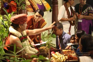 A photograph of a seated figure in an orange-red turban and orange cloth, wearing a thick white and green jasmine garland with red flowers, receiving blessings. A woman in a rust-brown sari with a long rudraksha mala and glasses extends her right hand over the head of a man in a blue checked shirt, who sits beside a small child in white. Behind them a woman in white with a rudraksha mala and another in a purple and green sari hold red fruit or offerings. In the foreground is a wooden box holding red fruit, gold jewellery and rudraksha beads, and green leaves frame the scene.