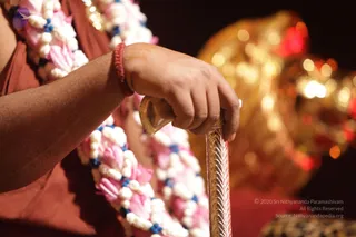 Close-up photograph of a person's right hand gripping the handle of a long ornate golden staff with engraved leaf patterns. The wearer's arm is covered in a rust-red or terracotta cloth, and a pink and white flower garland with blue beads hangs across the chest. A red thread is tied around the wrist. The background is blurred with warm golden and red lights reflecting off a large gold ornament.