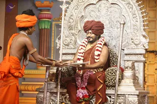A photograph of a seated man on an ornate silver throne with a carved lion-form armrest and leopard-print cushions. He wears a rust-brown turban, a brown robe with gold trim, and a white and red marigold garland with a purple flower garland across his lap, and holds a sheaf of papers. To the left, a standing attendant in an orange turban and orange cloth, wearing a pearl-bead necklace and pearl bracelets, hands him the papers. Behind them is a painted colourful pillar backdrop.