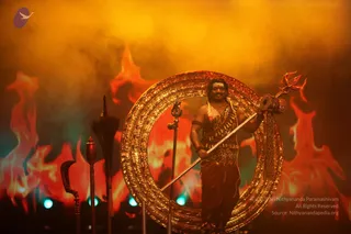 A figure stands on a stage within a large ornate golden ring, holding a long staff topped with a trident. He wears a leopard-print bodice with a dark red dhoti, a rudraksha-style mala and a tilak on his forehead. Behind him, orange and red flame-like backdrops and orange haze fill the stage, with teal stage lights at the base. To the left, a curved ceremonial dagger-like implement and a broom-like staff stand upright. The scene is lit as a dramatic stage performance.