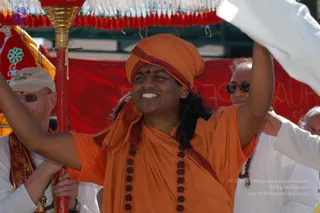 A smiling man in orange saffron robes and an orange turban with a red bindi stands under a red-tasselled canopy pole, holding it aloft with one raised arm during a procession. He wears a long dark rudraksha mala and a red-and-maroon sash over his shoulder. Behind him, a man in a white cap and sunglasses grips the red pole, and another man in sunglasses and white clothing holds the canopy. A large red banner with white lettering is visible in the background. The scene appears to be an outdoor street procession.