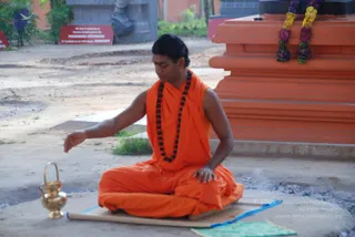 Outdoor photo of a man seated cross-legged on a mat in a saffron-orange robe with a dark rudraksha mala around his neck, eyes lowered and one hand extended over a small brass kalasha-style vessel with a handle on a stone surface. Behind him is a tall orange stone pedestal with purple and yellow flower garlands draped over it, and a red signboard and grey stone sculpture are visible in the background. A thin blue cord runs along the mat and a faint smoke haze rises from the ground.