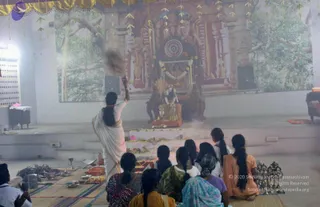 A photograph of a puja inside a hall with a large backdrop printed with a temple gopuram and tree foliage. A woman in a white sari with a long braid stands at left swinging a smoking incense or camphor vessel toward the altar. Seated devotees in front, several with long braids and dresses in orange, green, blue and purple, face the altar. The altar holds offerings on plates, a deity murti draped in garlands at the centre, and brass lamps and vessels on the floor in front. Smoke drifts through the air. Some people are visible at the left and right edges.