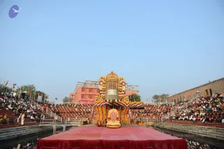 A wide outdoor photograph of a large amphitheatre-style stage with tiered seating packed with many spectators on both sides under a clear blue sky. In the centre stands a towering decorated lingam-form structure wrapped in colourful flower garlands and marigolds, set on an ornate golden base with a golden deity murti in front. Several figures in saffron and white clothing stand or sit around the base on a red-draped platform. Red-and-orange flags line the stairs and edges, and a red brick building stands behind the stage.