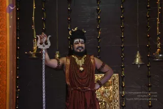 A standing man in a dark red-brown robe and gold-embroidered garment poses beside a tall silver trident held in his raised right hand. He wears a gold ornate necklace over multiple strands of rudraksha malas, rudraksha bracelets on his arms, a crescent moon ornament in his swept-up dark hair, a red bindi and white horizontal tilak on his forehead, and a full beard. His left hand rests on a golden ornamental plaque. Behind him are a dark stone wall hung with strings of lights, brass oil lamps and hanging brass bells. The scene suggests a darshan setting with a sacred Shaiva ornamental display.