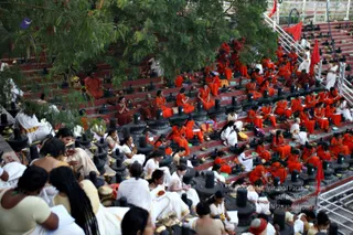 A large outdoor photograph of many people seated on red-painted stone steps lined with black Shiva lingams, performing a ritual. Most seated people wear saffron-orange robes, while others in white traditional clothes sit among them. Brass vessels and small oil lamps sit before the lingams, and red flags stand at the top of the steps beside green trees. Some participants hold open books or prayer items.