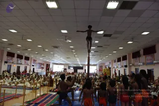 A large hall with white ceiling tiles, fluorescent lights and ceiling fans, lined with framed pictures and red curtains. In the centre, a shirtless young boy in black shorts balances with arms outstretched atop a tall wooden pole, while around him several men and boys in orange and red shorts or dhotis stand and crouch on blue mats and reach toward him. Seated devotees in white dhotis and saris fill the floor on the left and the rear, with striped rugs and brass stands in front. A golden figure of a deity and a kalasha-like arrangement are visible on a raised dais at the back. Some people sit in white on folded mats in front.