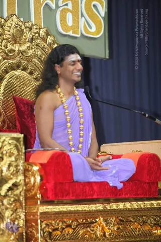 A seated man with long dark hair, white vertical forehead markings and a red dot, smiling with eyes closed. He wears a lavender wrap over bare shoulders and a long gold bead mala, with hands resting together in his lap. He sits on an ornate gold throne with red velvet cushions, a microphone stand beside him, and a blue-grey curtain behind. Partial letters on a green banner read 'ras' in gold glitter.
