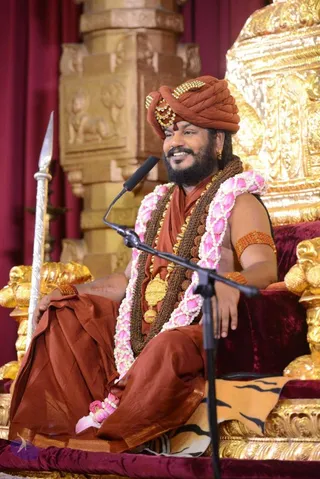 A bearded man in an orange-brown turban decorated with crystal and gold ornaments sits smiling on an ornate gold-and-crimson throne, speaking into a black microphone on a stand. He wears a rust-orange shawl over the shoulder, a dark brown rudraksha mala, a gold pendant, and multiple pink-and-white lotus garlands draped over his bare upper body. A silver spear rests beside him, and a tiger-striped cloth covers the seat. Behind him are carved gold pillars and maroon drapes. Given the appearance cues of a distinctive turban, rudraksha malas and ceremonial gold ornaments, this is a probable Swamiji seated in person giving a discourse.