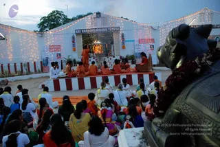 Evening scene in front of a white temple facade with string lights, a sign reading Sri Anandeshwara Temple above a shrine doorway with a lit deity murti inside. A group of devotees sits on a striped red-and-white platform and the ground facing the shrine; several men in orange robes sit in front, and a figure in orange stands at the shrine doorway. Women in white, yellow and blue saris and children in white and orange sit on the ground in rows. In the right foreground a dark bronze Nandi bull with garlands of red flowers faces the temple. Banners and framed portraits hang on the side walls, including a sign reading Kalpataru Darshan.