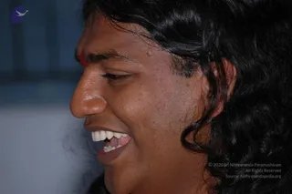 Close profile photograph of a smiling man with long, dark, wavy hair swept back past the ear. He has a red mark on his forehead between the brows and is laughing, showing his teeth. The background is a blurred blue-grey wall. Only the face and hair are visible; no clothing or setting details beyond the blur can be seen.