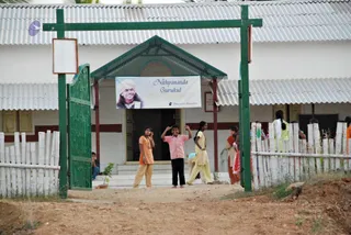 A photograph of the entrance to a white, tin-roofed building with a green wooden gate and white bamboo fence. Above the doorway hangs a banner reading Nithyananda Gurukul with a smiling portrait of a man wearing a white turban and dark clothing, and a small Nithyananda Foundation logo. Several children and young people stand around the entrance: a girl in an orange top and cream trousers, a boy in a pink shirt and black trousers raising his hands, a girl in a cream salwar kameez, and others in the background, including one in green. The ground is bare red earth.