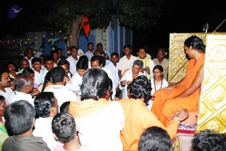 Night photo of a crowd gathered around a raised golden-panelled platform, where a figure in orange robes sits cross-legged on a red cushion at the right edge. A man in a white shirt and white dhoti kneels or bends toward the seated figure, and a person in orange garments leans in at the lower right. Many men in white shirts and dhotis press around, a few in striped shirts, and a woman in white with a red-beaded necklace stands near the platform. A strip of small coloured triangular flags and lights hangs on a blue-and-white building behind a tree, and a red-and-black flag is visible among the leaves.
