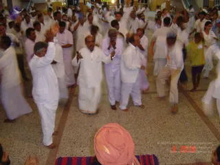 A dense indoor meditation gathering on a speckled terrazzo floor, with many participants dressed in white, moving with raised arms and hands clasped in namaste. Several men in white dhotis and shirts dance or sway in the foreground, while a few women and a child in a yellow-green top join in on the right. In the lower foreground, a figure seen from behind wears a pink head wrap and sits on a striped mat, facing the crowd.