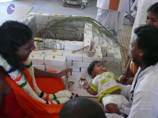 A photograph of a ritual at a brick-lined fire pit (homa or yagna) with a palm-frond arch and marigold petals scattered on the bricks. A long-haired figure in an orange-saffron shawl and a multicolour flower garland sits at left, extending a hand toward a baby in a white outfit with green and yellow floral trim who lies on a white cloth beside a small brass pot and a cup. A man in white shirt with a dark cord on his neck sits at right, a woman in a sari partly visible behind him, and a figure in white dhoti and orange sash stands at top. Incense sticks stand in a small bowl near the pit.