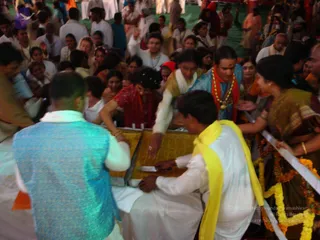 A crowded festive scene at a outdoor event with a green floor covering. In the centre, a young man in a turquoise sleeveless jacket over white clothes stands at a gold-draped table, while another man in white with a yellow shawl leans over it, handing or receiving a small paper slip. Behind the table, a man in a light blue top with a red-orange shawl and a long gold necklace stands in the crowd, and several women in saris, including one in dark green with gold border and another in maroon, reach toward the table amid garlands of orange and yellow marigolds. Many onlookers in white and colourful clothes press around.