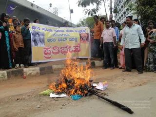 A street protest in an urban road beside an elevated overpass and tall buildings. In the foreground a small fire burns on the asphalt, consuming papers, a torn cloth and a charred wooden stick. Behind it, a yellow banner with red and black Kannada lettering is held up by several people; the banner shows two portrait photographs, one of a woman with long hair and one of a smiling man in an orange turban. A man in an orange shirt and another in a pink-and-white striped shirt stand near the fire, and a man in a light blue shirt and dark trousers stands at right. Women in dark dresses and salwar kameez watch from the left and right, and children stand at the left edge.