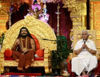 Two men sit on ornate gold thrones on a stage decorated with gilded carved panels, golden lamps and green foliage. On the left, a man with long dark hair, a beard and a saffron-brown robe with a gold-beaded necklace and a rudraksha mala sits with a bracelet on his wrist, smiling and gesturing. On the right, an older man with white hair wears an all-white dhoti and shirt, holds a microphone in both hands and speaks. Behind them, a painted figure in a pink turban and a deity murti in pink and purple stand on the dais. A microphone stand is positioned in front of the thrones, and a basket of fruit sits at the lower right.
