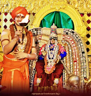 A young priest in saffron-orange robes and an orange turban holds a lit oil lamp (aarti) toward a deity murti seated on an ornate golden throne. The priest wears wooden rudraksha malas, a gold chain necklace, a bead armband and white sacred markings on his arms and forehead. The deity murti is clad in a blue blouse and maroon skirt, with a jeweled crown, layered gold and pearl necklaces and a garland of orange and pink flowers, and holds a golden hand gesture. Behind the throne hang a green velvet curtain and gold-embossed panels with red diamond cutouts. A brass conch shell sits at the base of the throne. Visible text in the lower corner reads Rajarajeshwari Brahmotsava Day 3.