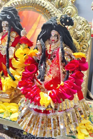 Close view of two deity murti figures on an ornate gilded shrine with a golden arched backdrop. The foreground figure has long black matted hair, a red sari with gold sequin embellishment and a pearl and red stone necklace, holding a trident-like implement in one hand. Garlands of red and pink rose petals and yellow flowers surround the figures. A second murti with similar black hair and red attire stands partly visible at the left. No physical people are present.