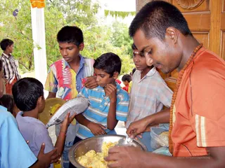Photo of a food distribution at a temple-style doorway. A man in an orange short-sleeved shirt with a rudraksha mala serves rice from a large steel bowl. Several boys crowd forward to receive food; one in a blue striped polo holds a plastic bag, another in a plaid shirt has a tilak on his forehead, and a boy in a striped shirt stands at the front. Green leaves and an orange cloth hang above, with trees behind. Trees and a light-coloured pillar frame the scene. No a person candidate is clearly identifiable from appearance.