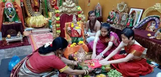 Indoor temple puja in a shrine room decorated with gold-framed pictures, flower garlands and small deity figures. A seated priest in white dhoti and a rudraksha-style mala watches from the back. Three women in saris sit on the floor around a brass yantra or small temple structure and a red cloth with offerings: a bowl of kumkum, fruits, grapes and flowers. The woman on the left wears a red blouse and grey-silver sari; the centre woman wears a magenta sari; the woman on the right wears a red sari. A large seated deity murti with a red and orange garment and yellow flower garlands is at the centre, with oil lamps nearby.