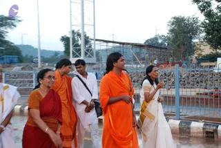 A photograph of a group walking outdoors along a wet paved path beside a blue metal railing, with a tall lattice telecom tower and wind turbines in the background. A man in the centre wears an orange-saffron shawl over his shoulder and a gold-bordered orange cloth, with long swept-back dark hair, and gestures while talking. Beside him a woman in a white sari with gold border holds a phone and walks with a woman in a red sari and orange blouse on the left. Behind them a younger man in an orange cloth and another in white shirt and trousers with a shoulder bag walk along. Bamboo scaffolding and stacked stones are visible on the right. Swamiji (probable): the central man has long swept-back hair and saffron-orange ornate clothing, consistent with the page description.