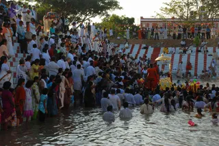 A large crowd stands and wades in a river at dusk, with many people in white and cream clothing and women in colourful saris and salwar kameez on the near bank. On the right, a palanquin-style golden canopy with a gilded seat and yellow-orange drape sits at the water's edge, attended by a figure in saffron robes. Red-and-white stepped ghats rise behind the crowd, with drummers and onlookers in white along the top steps and red flags and bunting strung across the grounds. Colourful balloons float on the water near the palanquin, and a pink balloon drifts in the foreground.