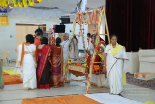 A photograph inside a hall decorated with yellow and orange marigold garlands. A wooden swing frame draped with orange and yellow flower garlands and a cushioned seat stands at centre. Several people in white and orange clothing gather around it: a woman in white with an orange shawl at left, women in red, maroon and orange saris beside the swing, and a man in white with a lanyard behind it. On the right, a woman in a white sari with a yellow shawl walks past. Orange and yellow floor cloths lie on the floor, a cushioned sofa sits at right, and a maroon curtain hangs in the background.