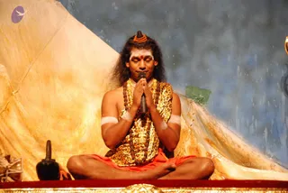 A photograph of a man seated cross-legged on a raised golden stage with eyes closed and palms pressed together in namaskar around a black microphone. He has long dark hair swept back, a rudraksha-style bead crown on his head, white horizontal tilak lines and a red dot on his forehead, bare upper arms with white bands, and a leopard-print shawl over bare shoulders. Layered rudraksha malas and a gold ornamental necklace hang on his chest, with a saffron-orange dhoti. Behind him are gold and golden-yellow draped cloths and a painted blue backdrop with a green patch. A black pot sits at left on the stage edge. Appearance cues are distinctive (long hair, crown, rudraksha malas, ornate shawl), so this is an appearance-based probable candidate for a person.